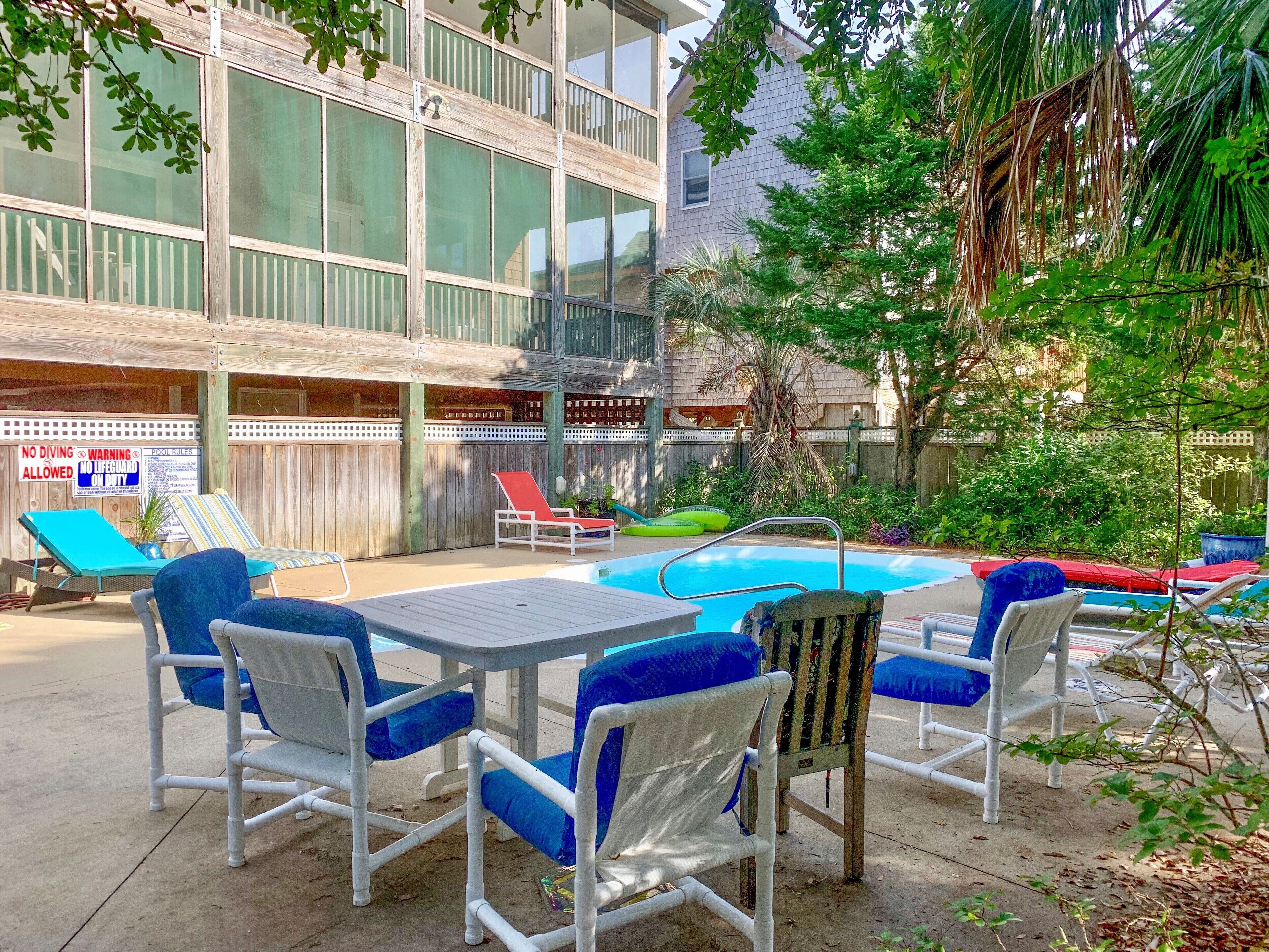 An outdoor pool area with lounge chairs and a table, surrounded by a wooden fence and greenery.