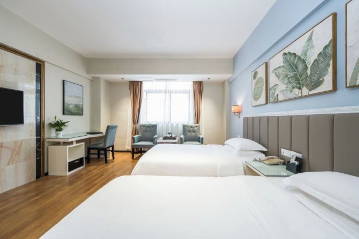 Quarto de hotel com duas camas, uma escrivaninha, uma cadeira, uma televisão e obras de arte na parede.