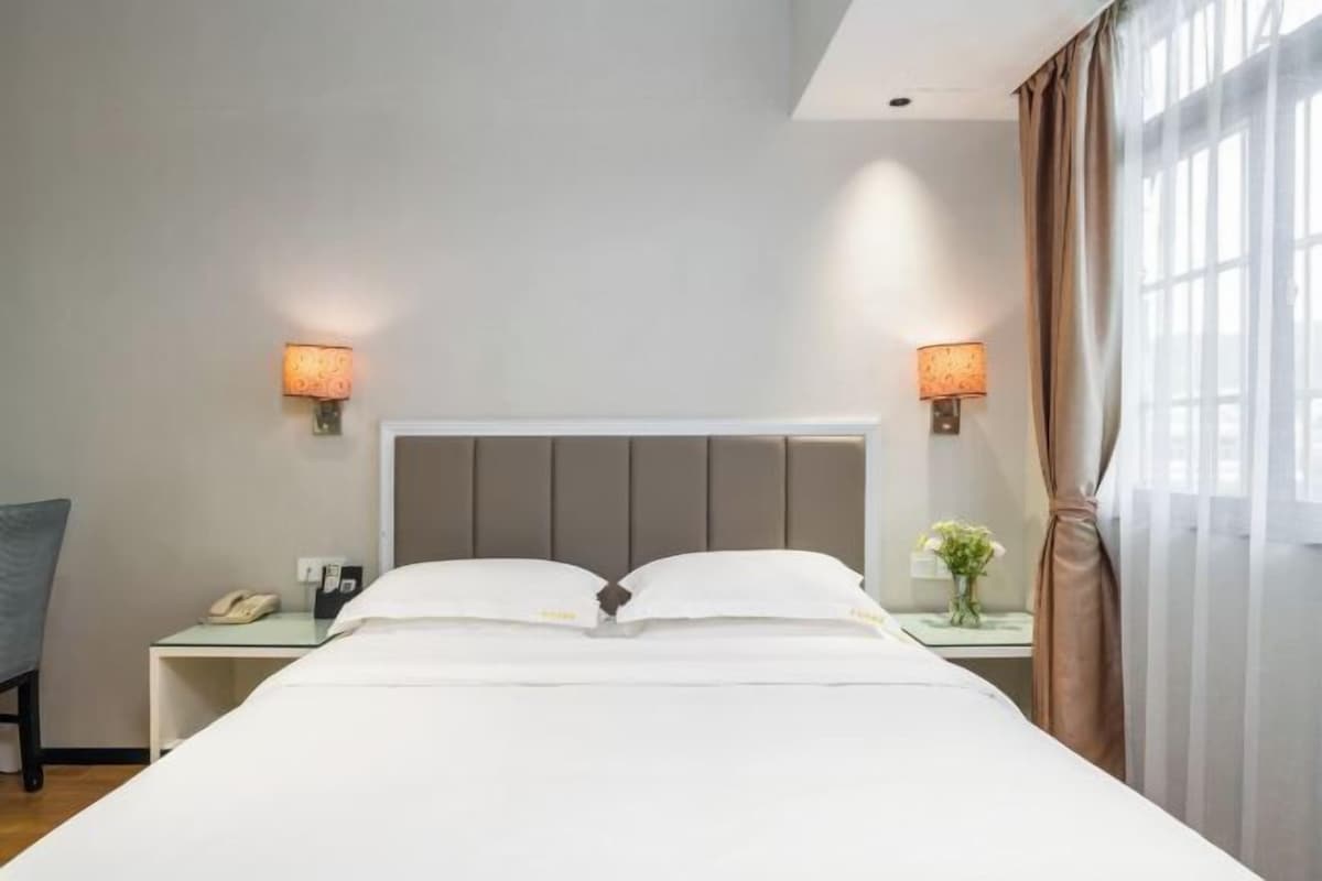 A neatly made bed with white linens, two bedside lamps, a nightstand with a phone and a vase of flowers, and a window with curtains.