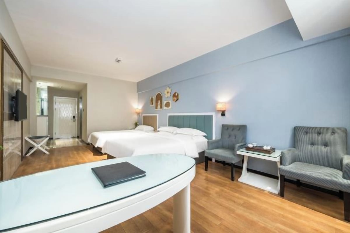 Une chambre d’hôtel avec deux lits, un coin salon comprenant une chaise et une petite table, une table en verre avec un livre, et un mur décoré de différents objets.