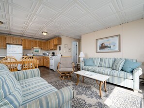 A living room with a striped sofa, a rocking chair, a coffee table, and a dining area with wooden chairs and a table. - Oceanfront condos with rare 2 night minimum rentals and linens included! (Ocean City)