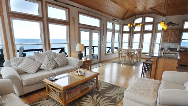 A spacious living room with a large window overlooking the sea, a cozy seating area, and a kitchen area. - Hooligan Sunset:  Waterfront deck, sunsets, private boat dock. (Ocracoke)