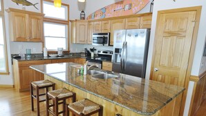 A kitchen with wooden cabinets, a stainless steel refrigerator, a granite countertop, and four bar stools. - Hooligan Sunset:  Waterfront deck, sunsets, private boat dock. (Ocracoke)