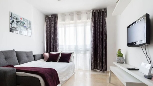 Un dormitorio moderno con una cama, un sofá, un televisor y un ventanal con cortinas.