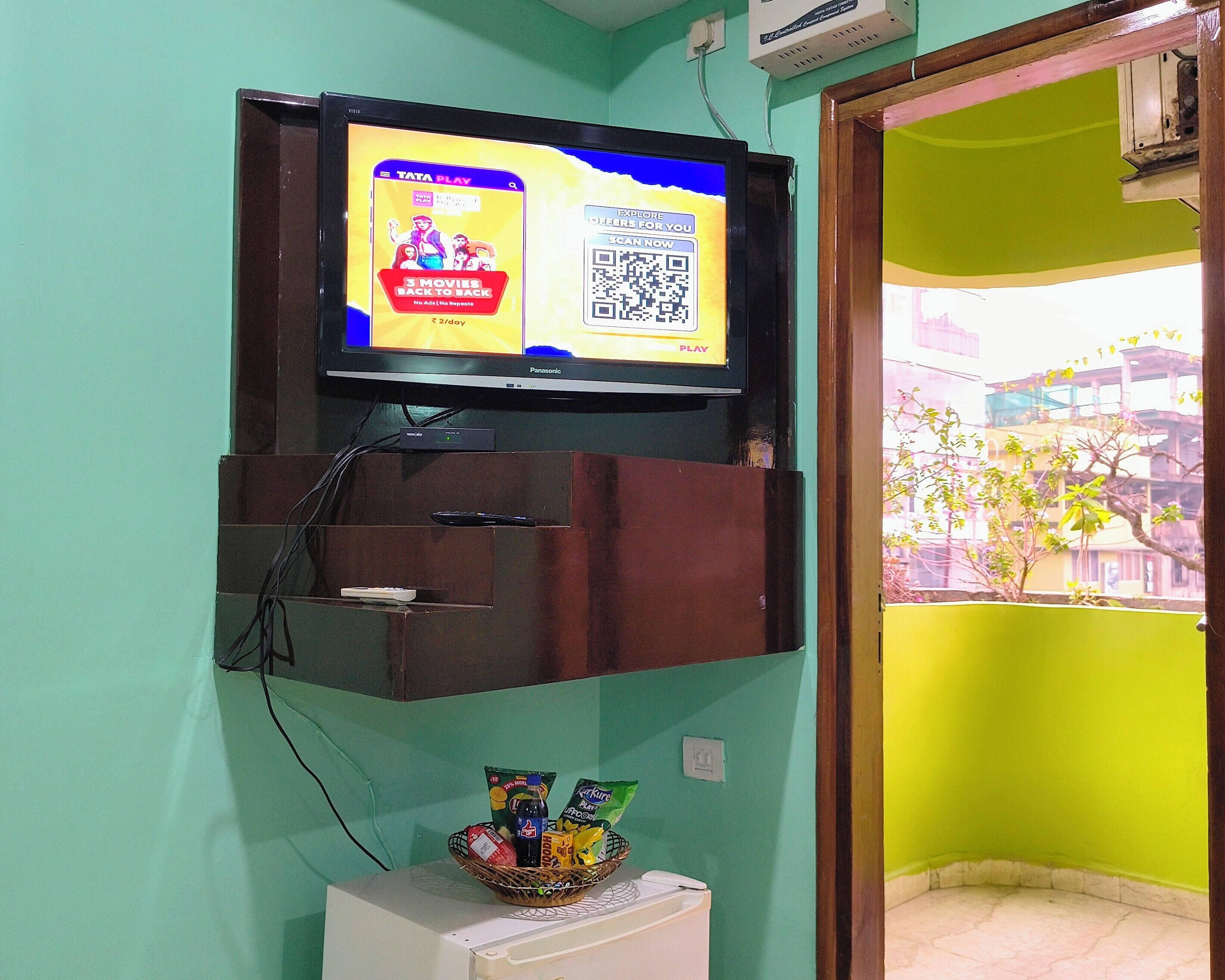 A television mounted on a wall with a QR code, a basket with snacks, and a white appliance below it.