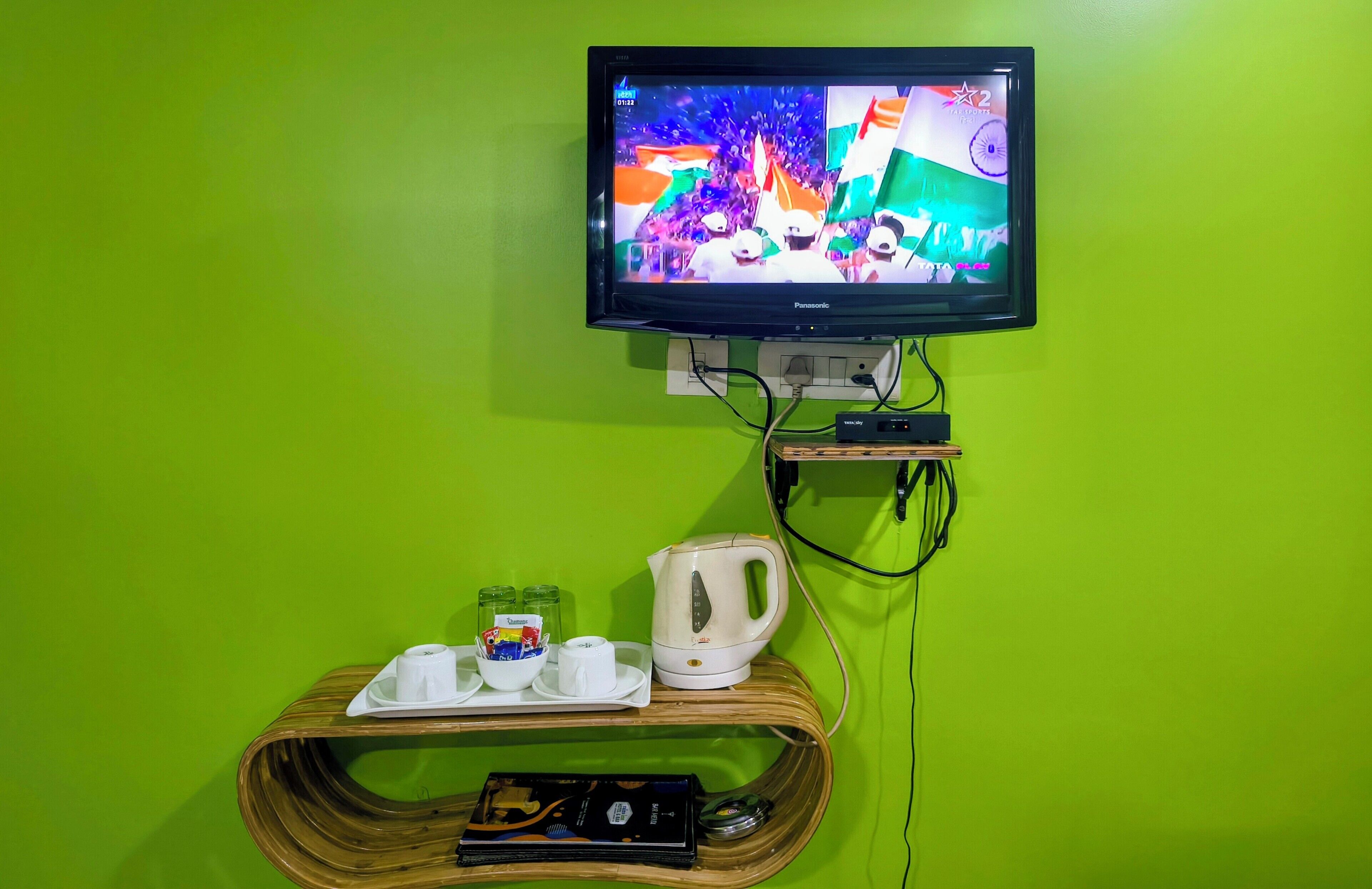 A television mounted on a green wall, a wooden shelf with a kettle, cups, and snacks, and a power strip.