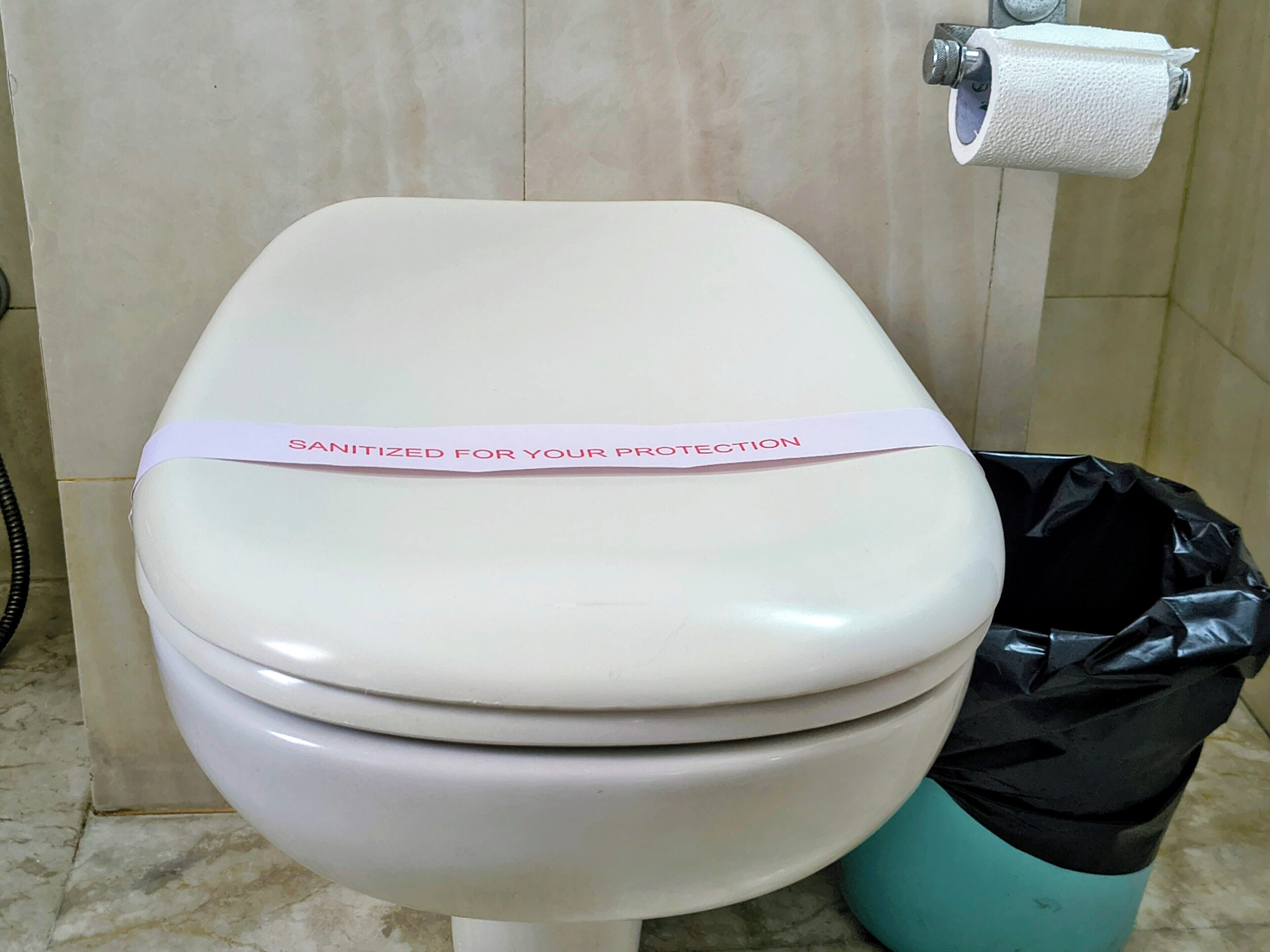 A toilet with a 'SANITIZED FOR YOUR PROTECTION' label, a toilet paper holder, and a black trash bag.