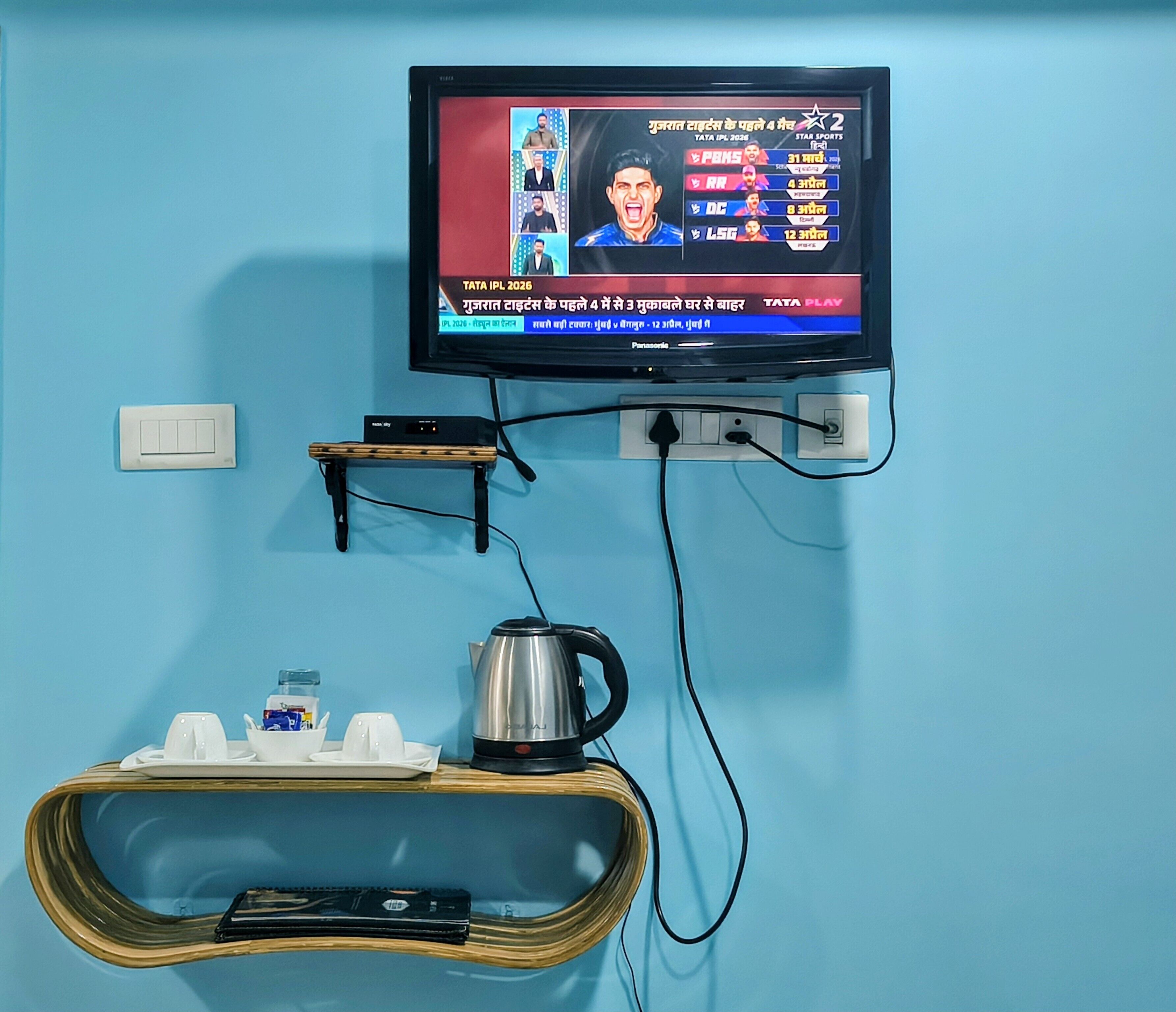 Un téléviseur fixé au mur, diffusant un événement sportif, une prise de courant, un interrupteur, une petite étagère avec une bouilloire et des tasses, et un câble relié au téléviseur.