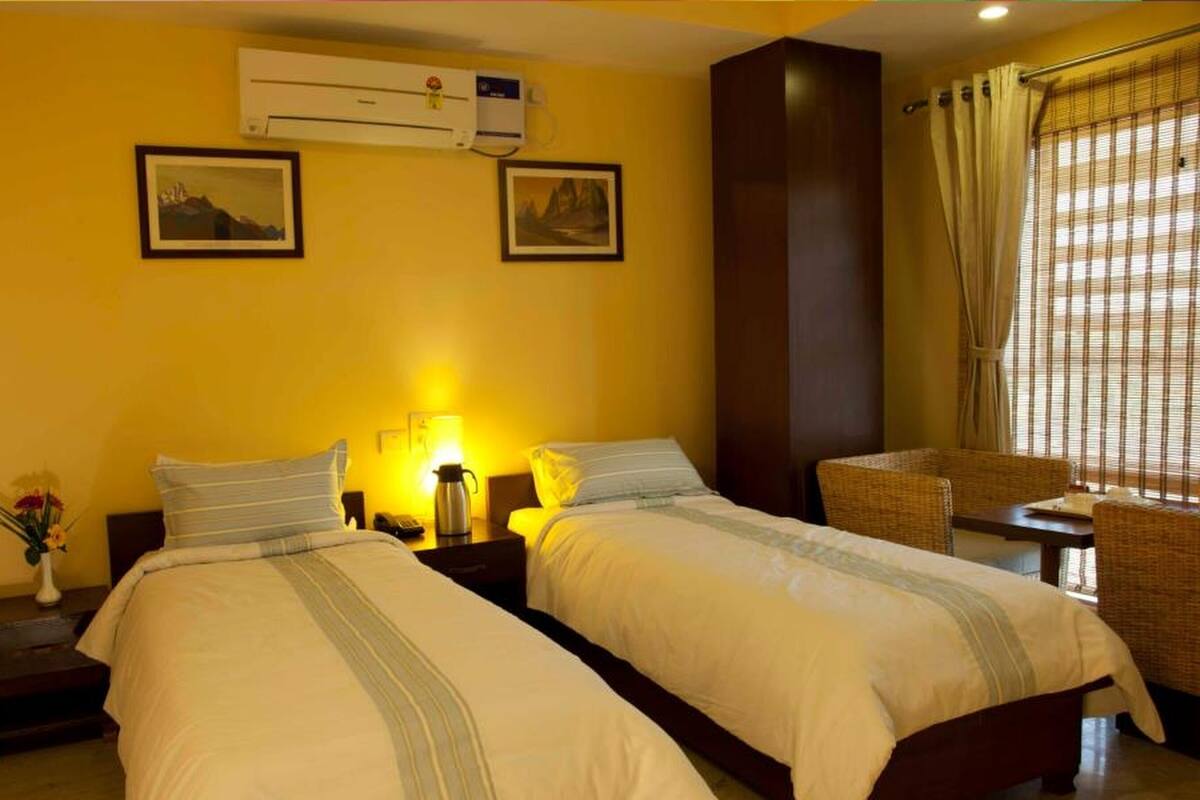 A hotel room with two beds, a nightstand with a lamp, an air conditioning unit, and a window with curtains.
