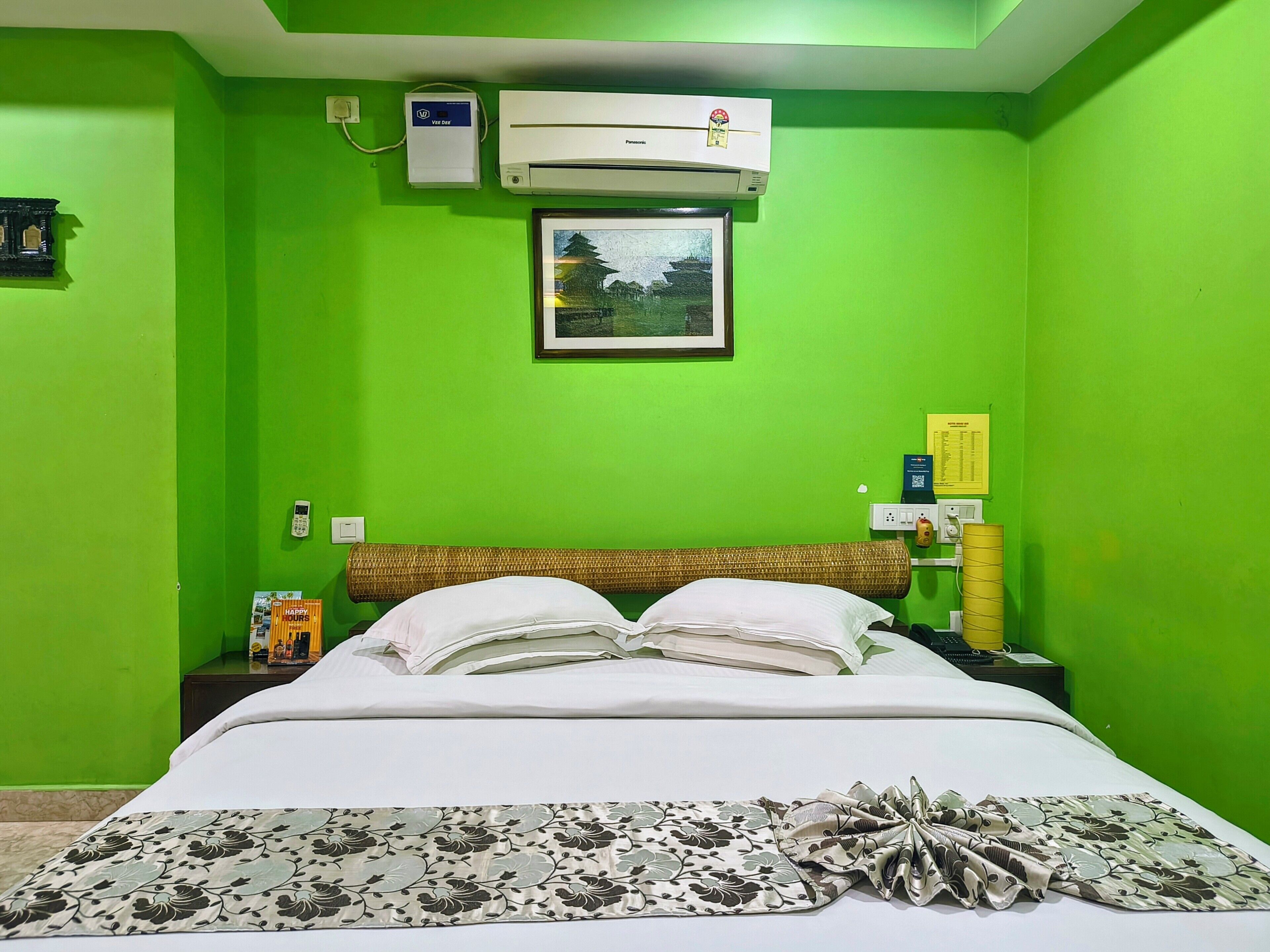 A room with green walls, a bed with white bedding, a picture on the wall, and an air conditioning unit.