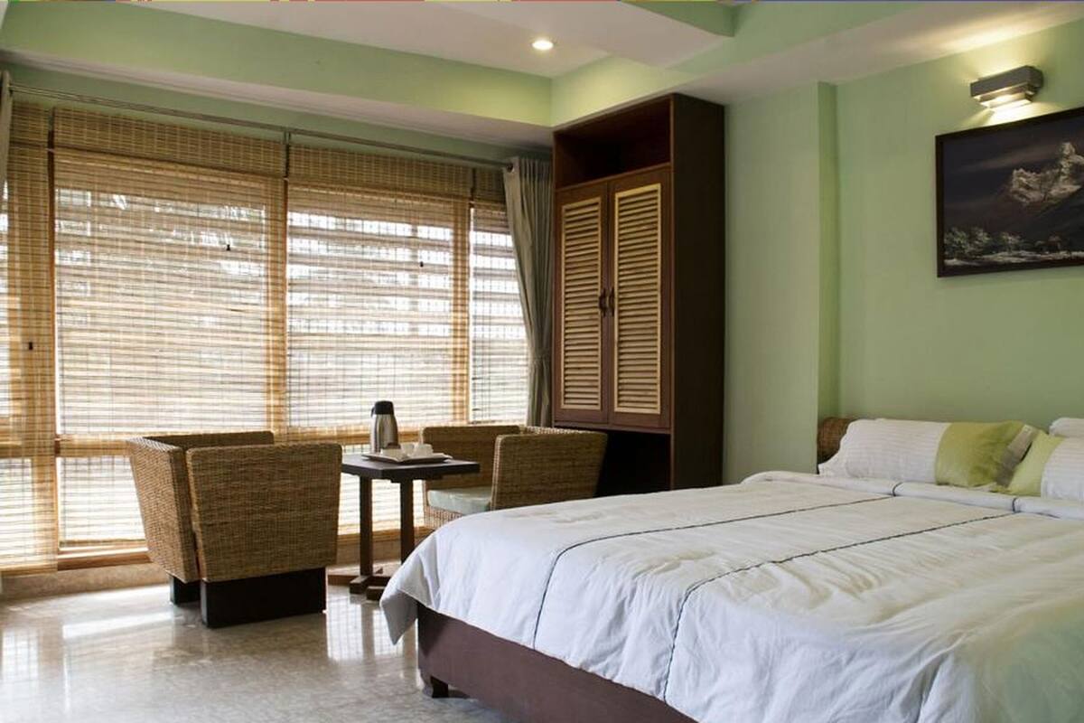A hotel room with a bed, a table with chairs, a painting on the wall, and large windows with blinds.