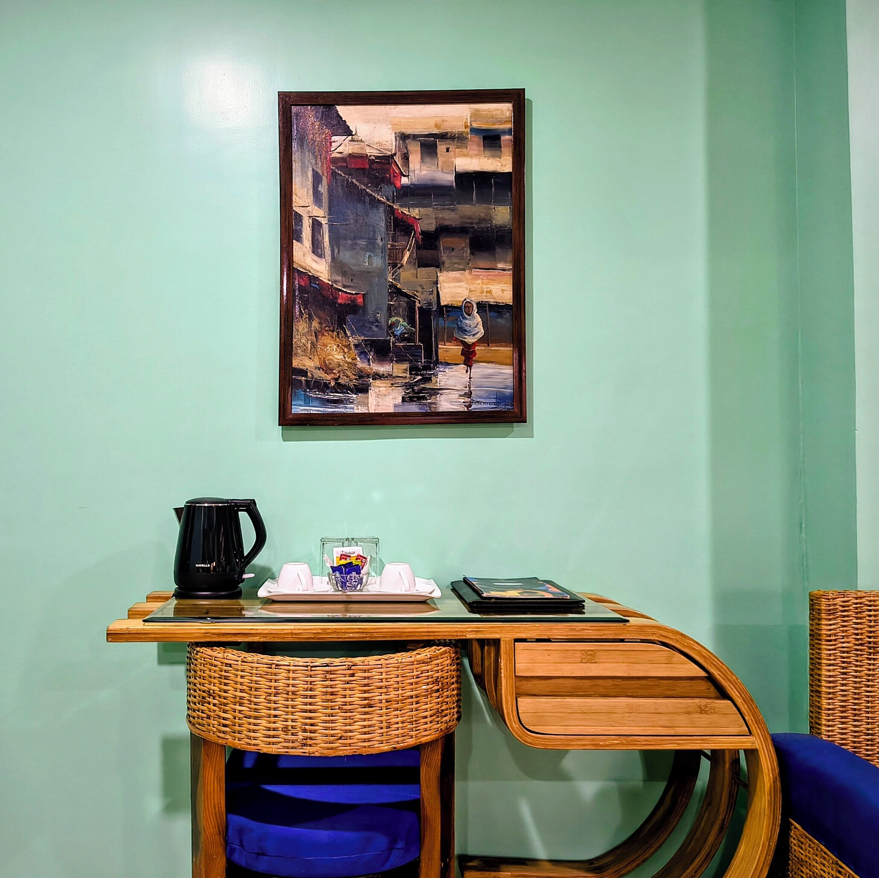 A small room with a painting of a street scene, a table with a kettle, cups, and a tray, and wicker furniture.