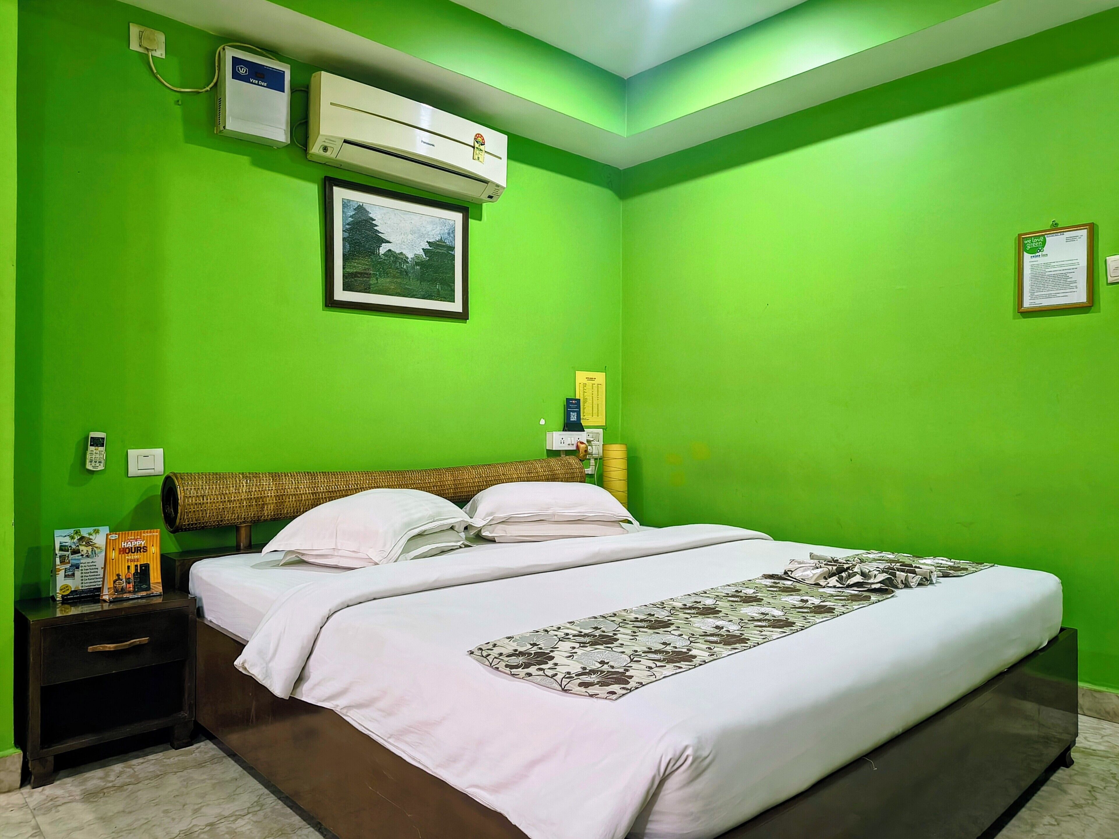 Une chambre d’hôtel avec un lit, une table de chevet, un climatiseur fixé au mur, un tableau encadré et un mur vert.