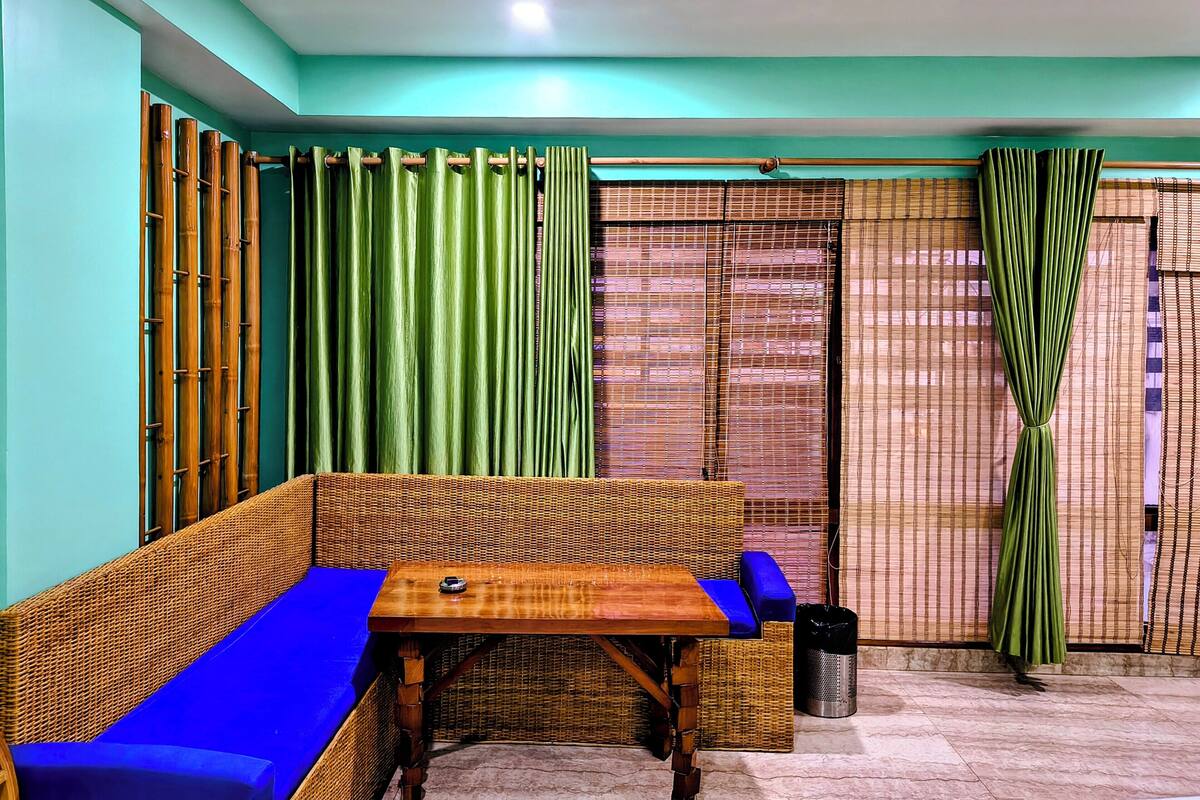 A room with a wicker sofa, a small table, a trash bin, and a view through a window with bamboo curtains.