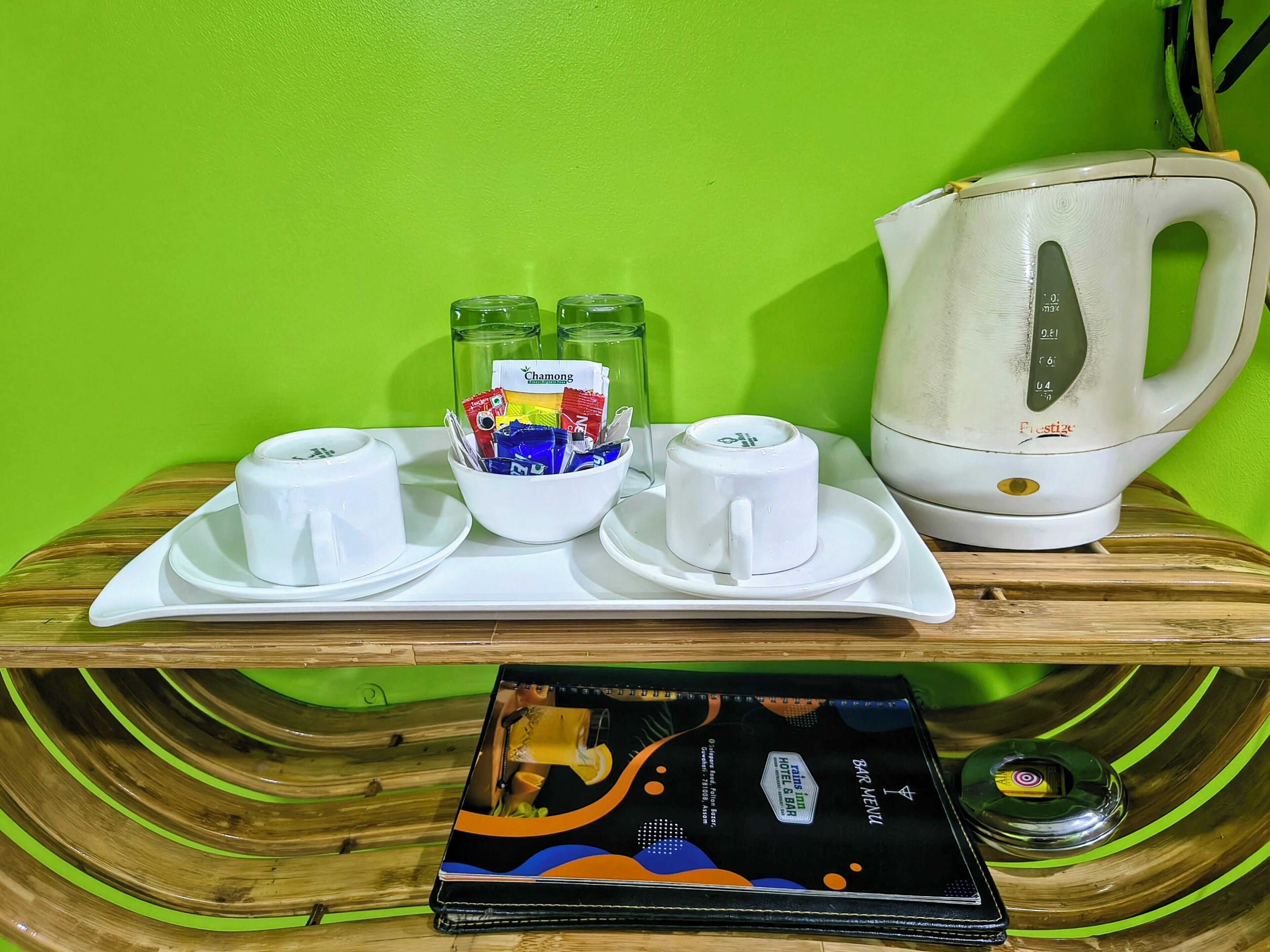 A breakfast tray with cups, a bowl of tea bags, and a kettle on a wooden table against a green wall.