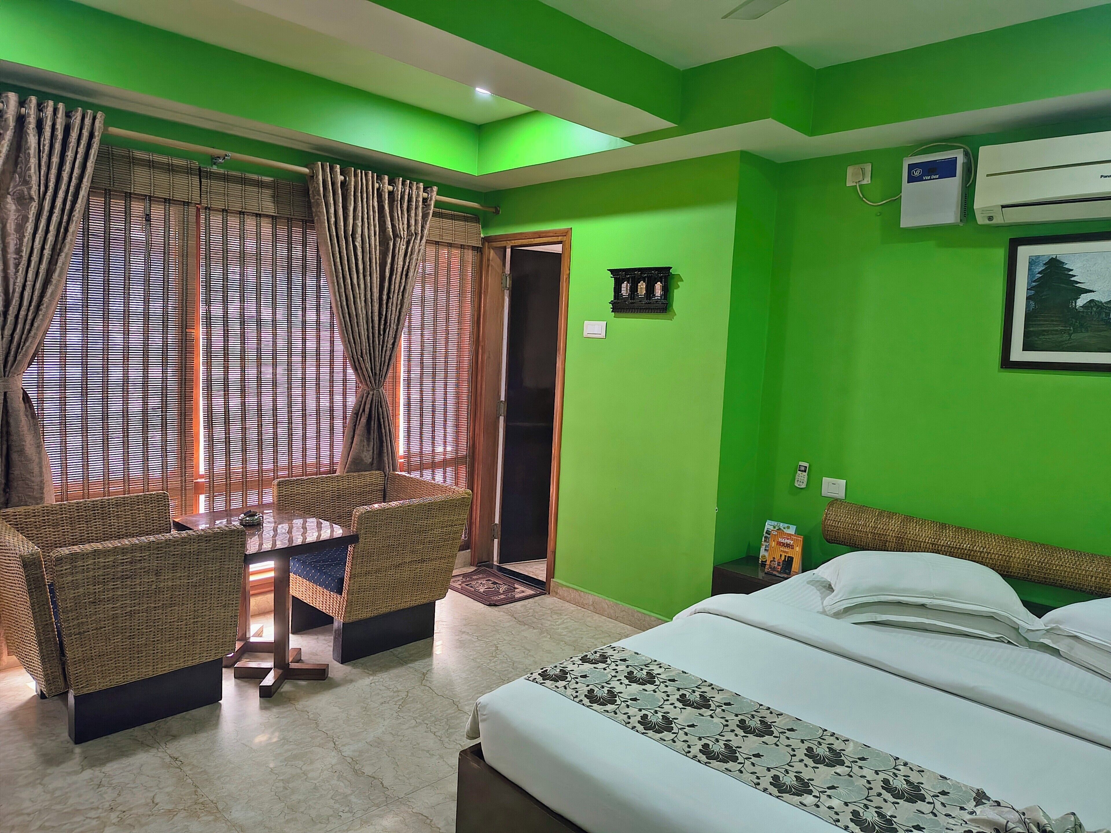 A hotel room with a bed, a small table, wicker furniture, a green wall, and an air conditioning unit.