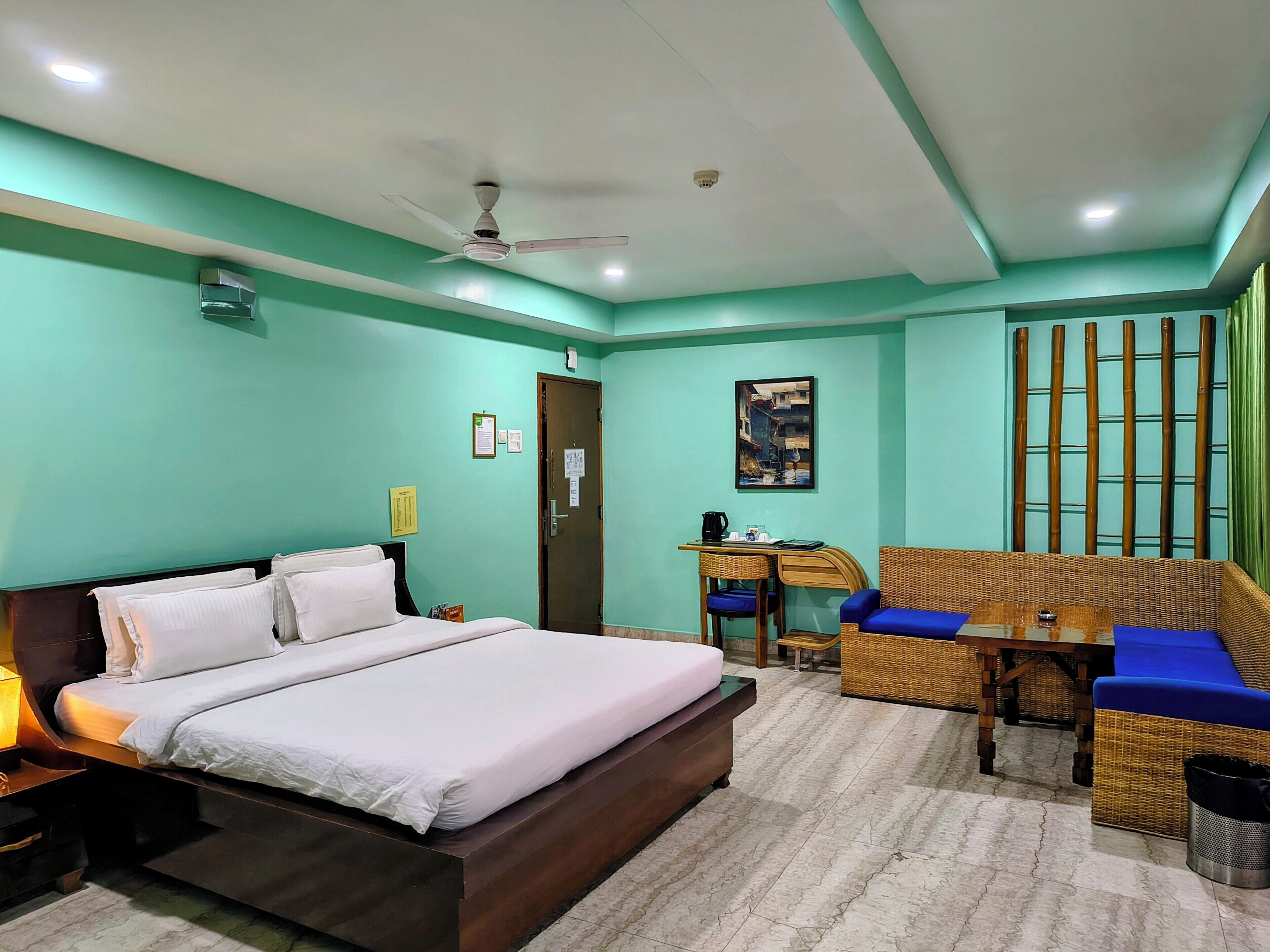 A hotel room with a bed, a desk, a chair, a sofa, a ceiling fan, and a wall-mounted air conditioner.