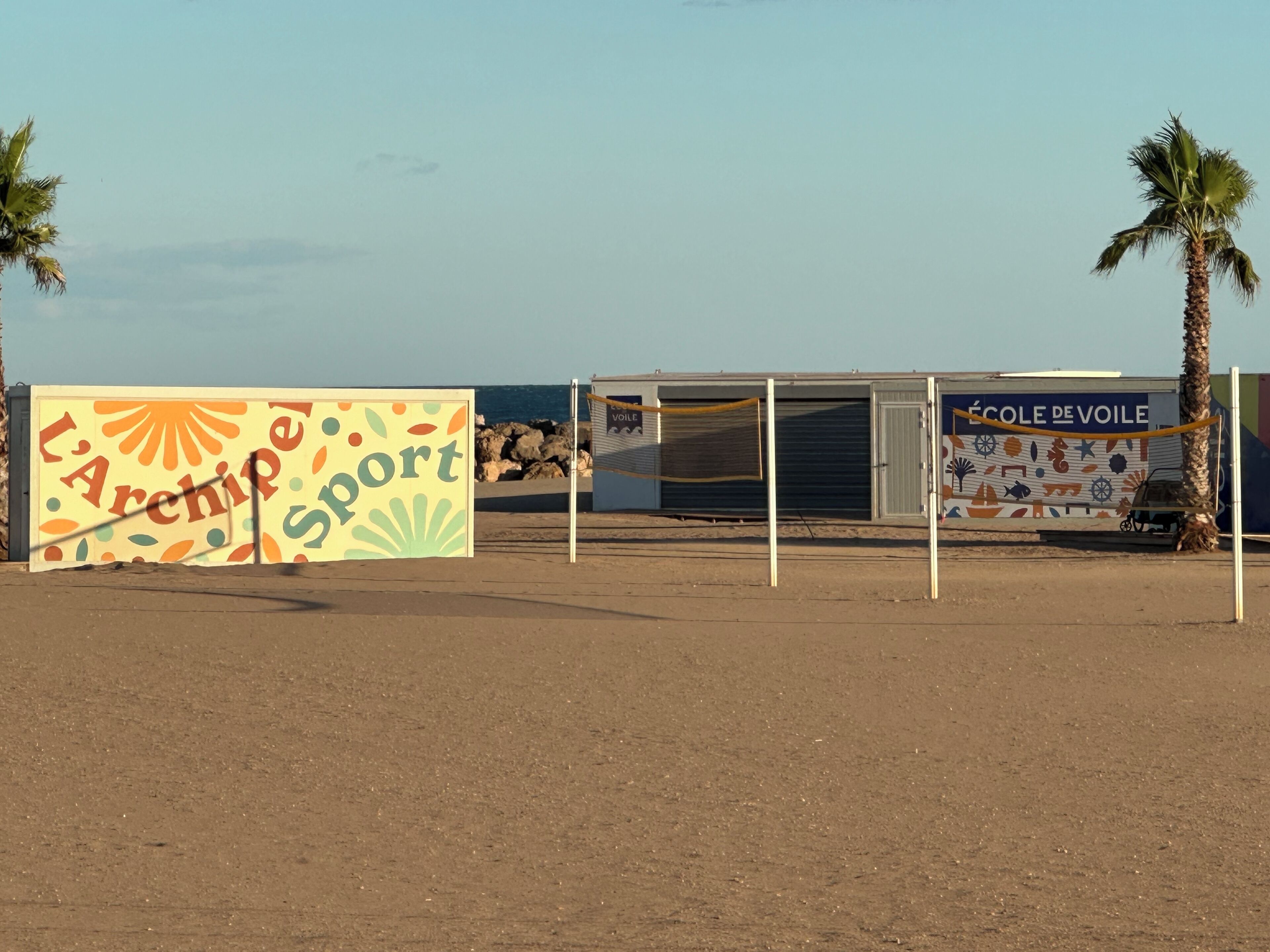 A beachside building with a colorful sign reading "L'Archipe Sport" and another sign for "Ecole de Voile".