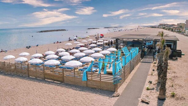 A beach with rows of umbrellas and sun loungers, a wooden fence, and a building with a sign. - Villa T3, private terrace & parking. Wifi Fibre, Swimming pool, Shops, Beach 50m! (Valras-Plage)