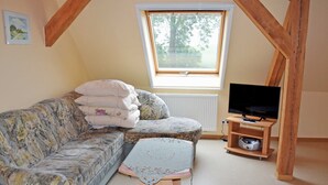 Een gezellige kamer met een bank, een klein tafeltje, een televisie op een houten standaard, en een raam met uitzicht op bomen.