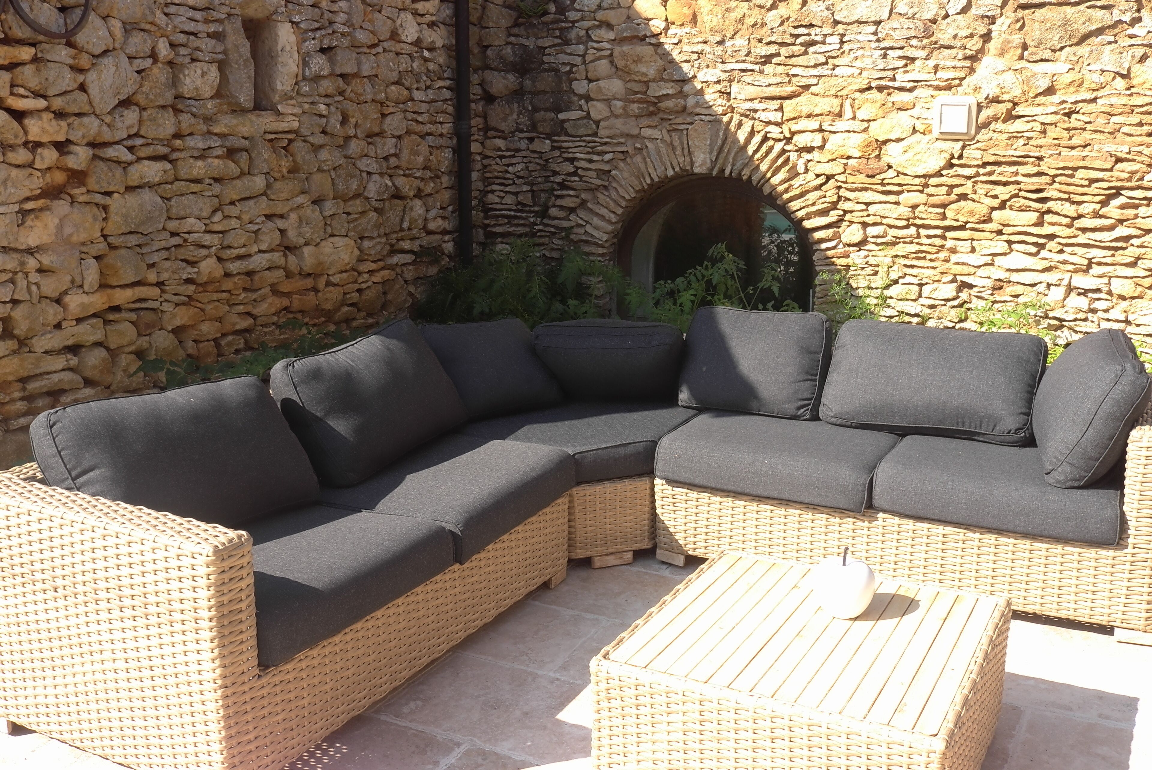 A wicker sectional sofa with dark cushions, a wicker coffee table, and a stone wall with a circular archway.