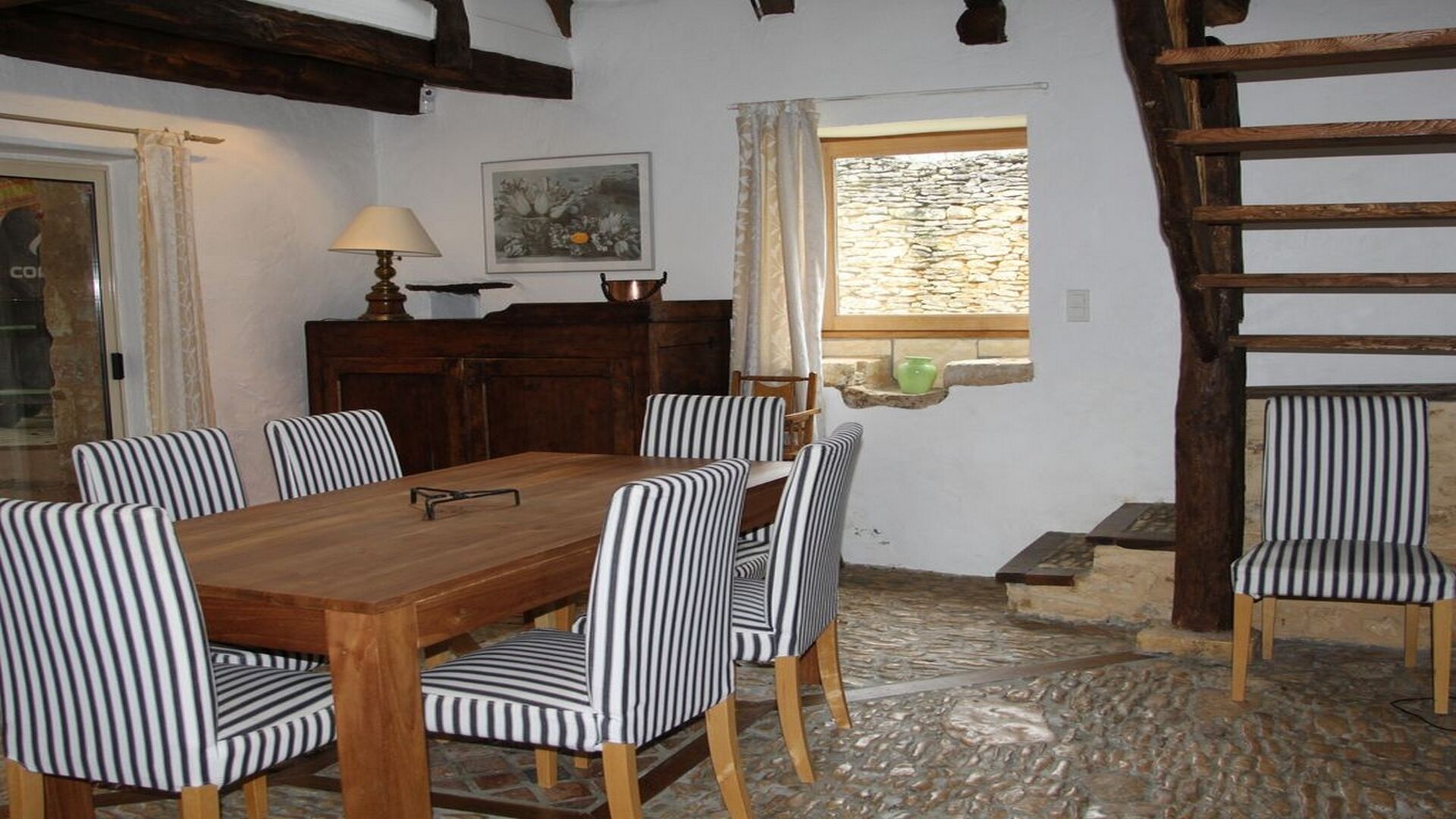 Un coin repas avec une table en bois et des chaises à rayures, une lampe et une fenêtre donnant sur un mur en pierre.