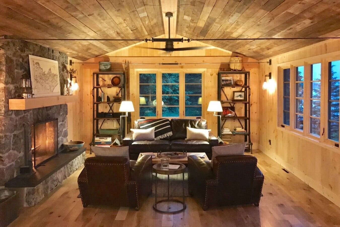 A cozy cabin interior with a stone fireplace, wooden walls, a leather sofa, and a bookshelf.