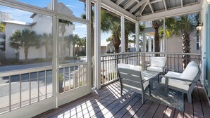A covered outdoor area with a wooden deck, white outdoor furniture, and large windows offering views of palm trees and buildings. - Seacrest Beach Home + Dog Friendly, Tram & Bikes (Seacrest Beach)
