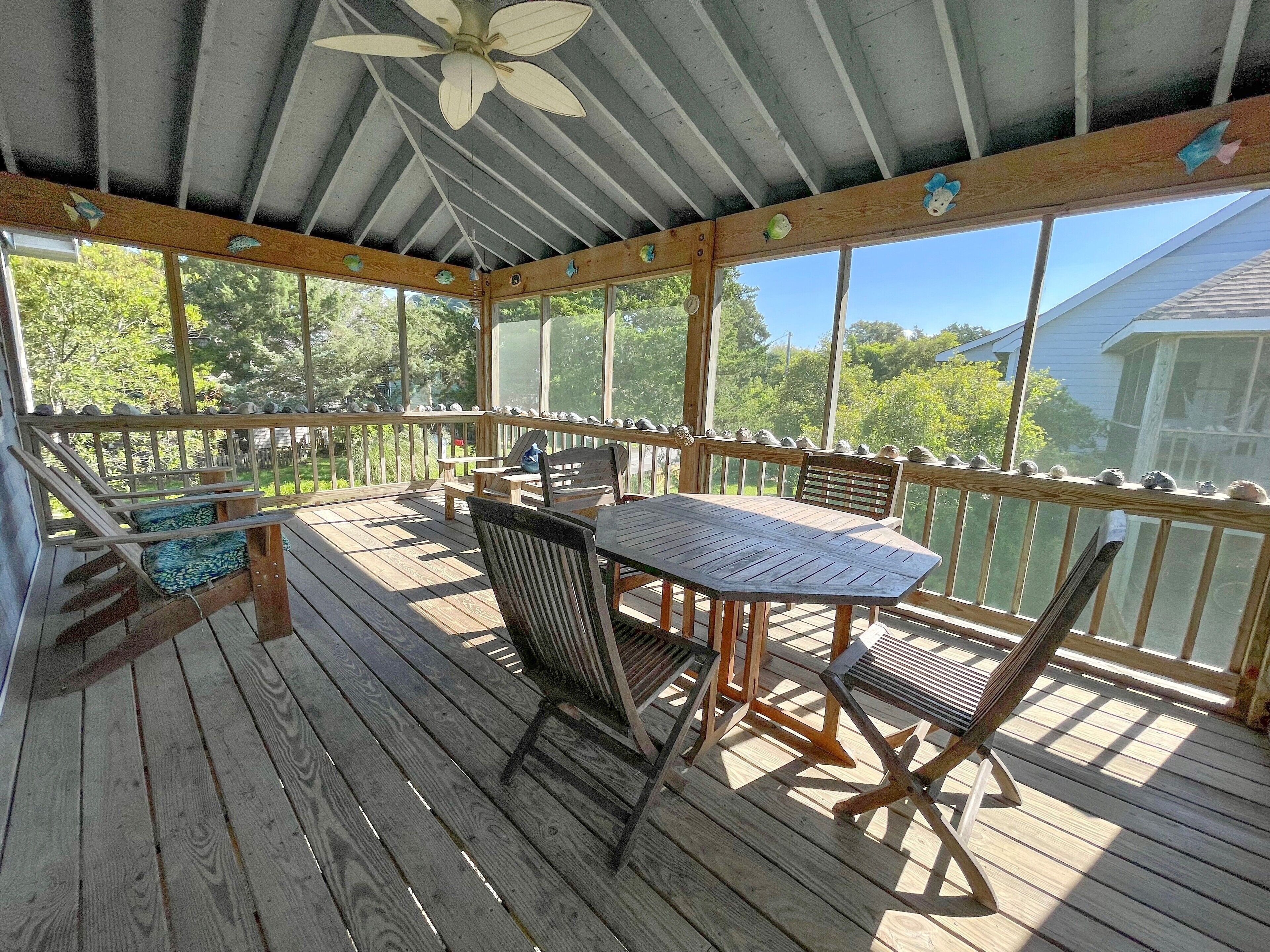 Una terrazza in legno coperta con tavolo e sedie, una ventola a soffitto e vista su alberi e una casa.