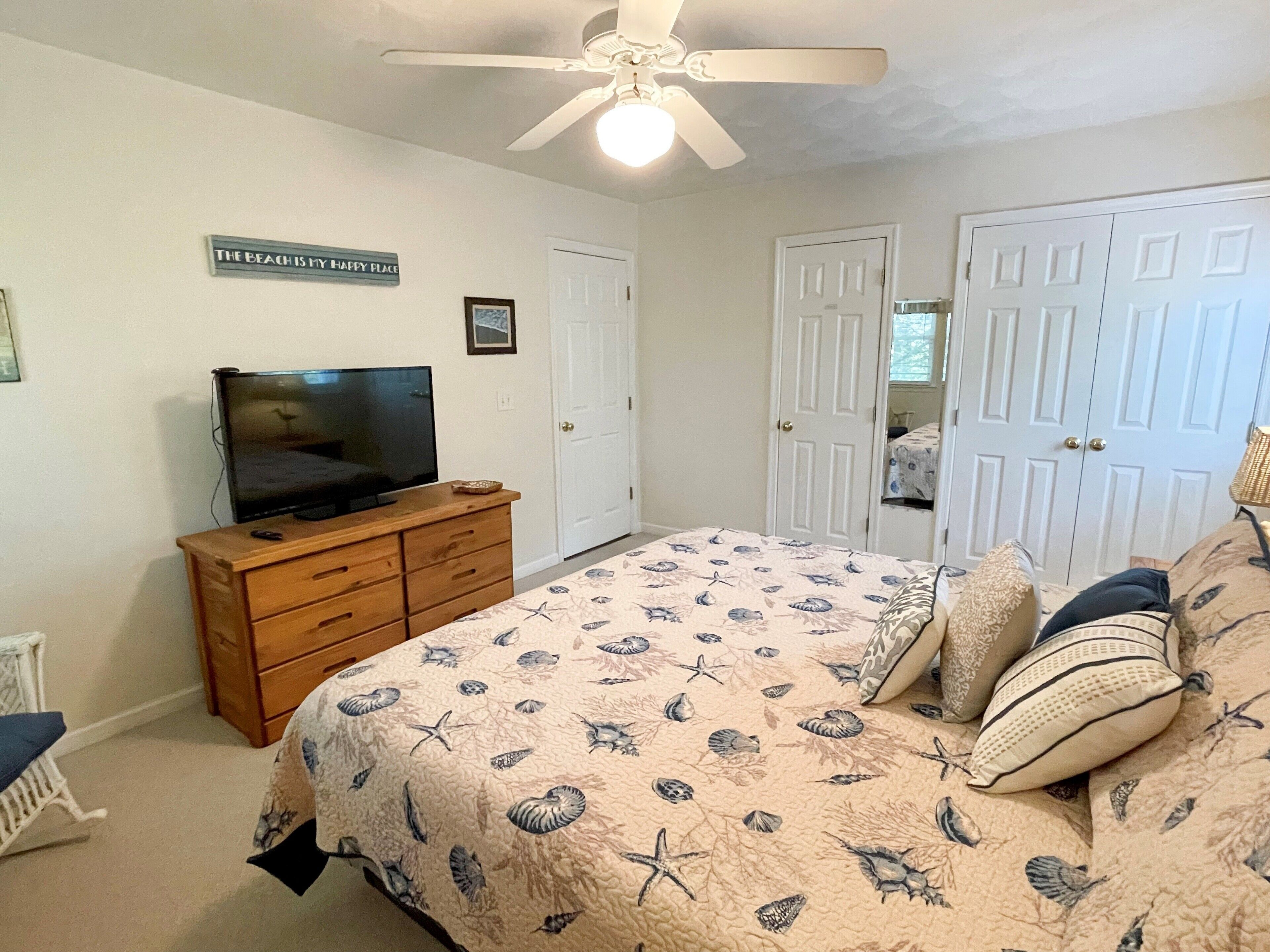 A bedroom with a bed, a television on a wooden dresser, a ceiling fan, and white doors.