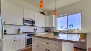 A modern kitchen with white cabinetry, a central island, and a large window with a view of palm trees. - Spanish Town home Just Blocks To The Beach With Panoramic Ocean Views (Oceanside)