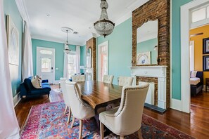 A dining room with a wooden table, white upholstered chairs, a chandelier, and a fireplace.