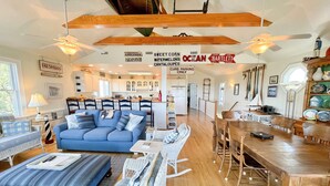 A spacious living area with a blue sofa, white wicker chairs, a wooden dining table, and a kitchen in the background. - Sea View: Rooftop deck with spectacular views. (Ocracoke)
