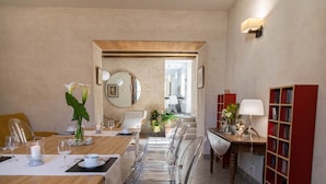 Un comedor con una mesa larga, sillas transparentes y una mesita de madera con una lámpara. Hay estanterías a la derecha y un espejo en la pared.