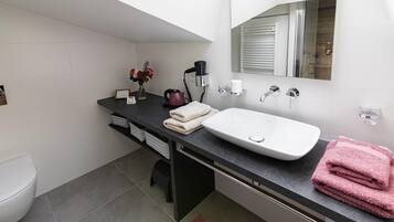 A modern bathroom with a white sink, black countertop, and a mirror above it. There are towels and a small vase with flowers on the countertop.