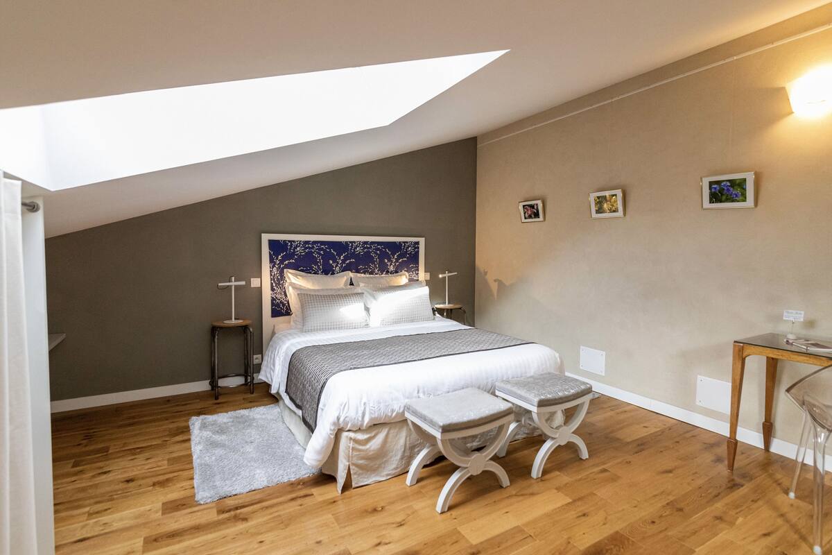 A bedroom with a sloped ceiling, a bed with white bedding, a small bedside table, a chair, and a desk with a chair.
