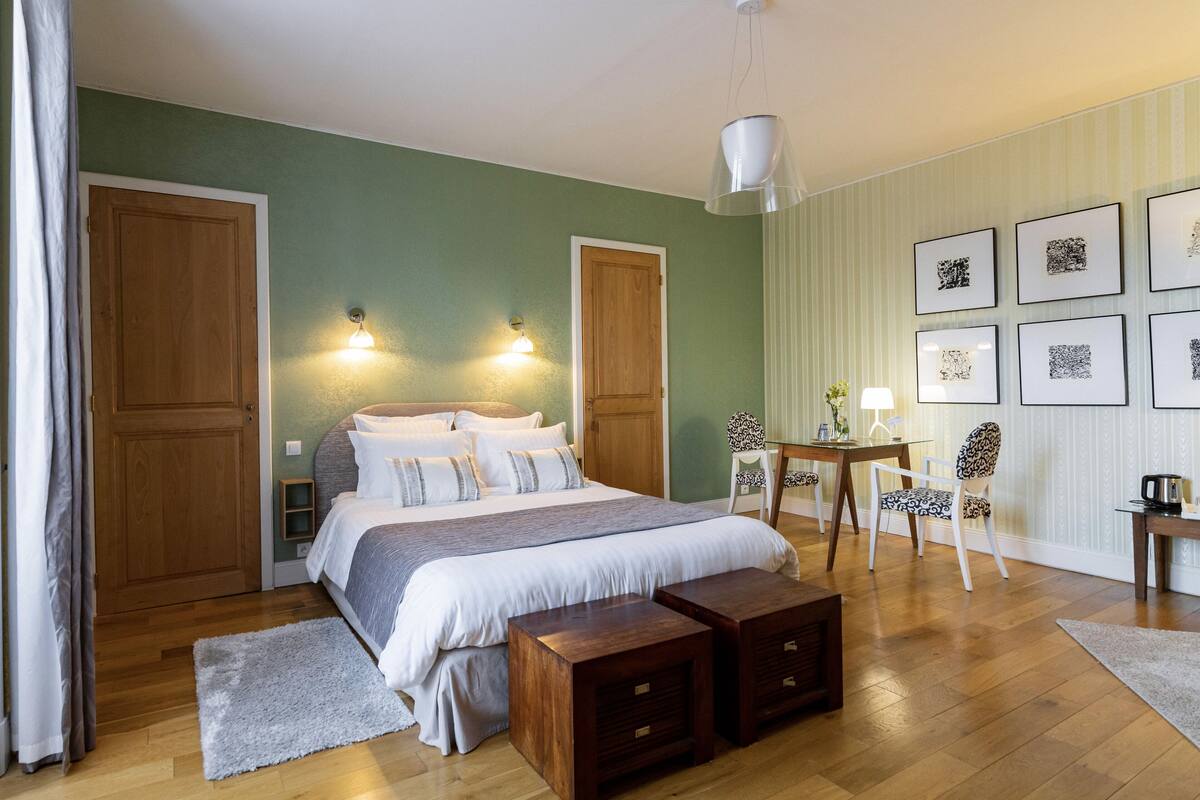 A bedroom with a large bed, two bedside tables, a desk with chairs, and artwork on the wall.