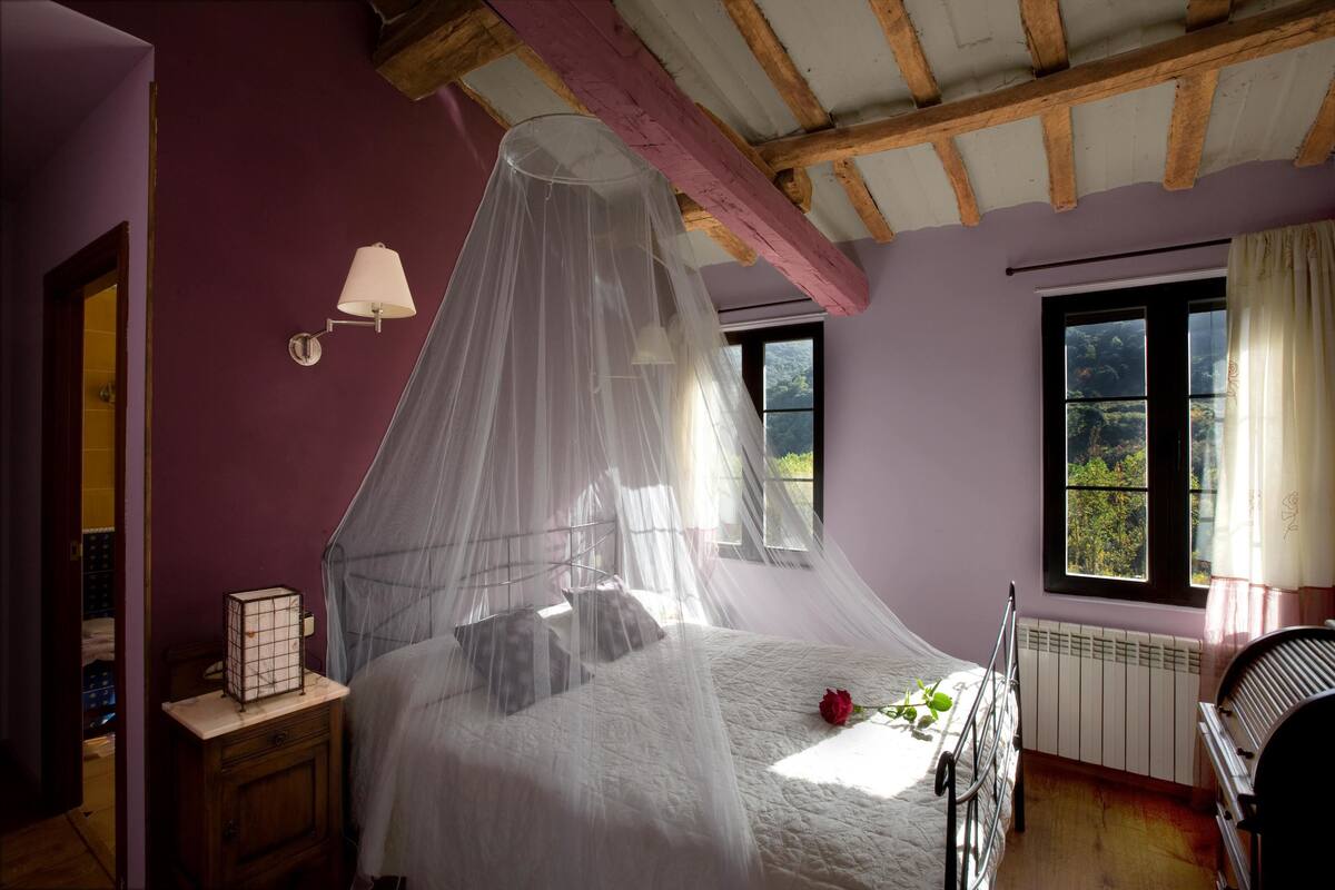 Un dormitorio con cama bajo dosel, mesita de noche, radiador y ventana con cortinas.