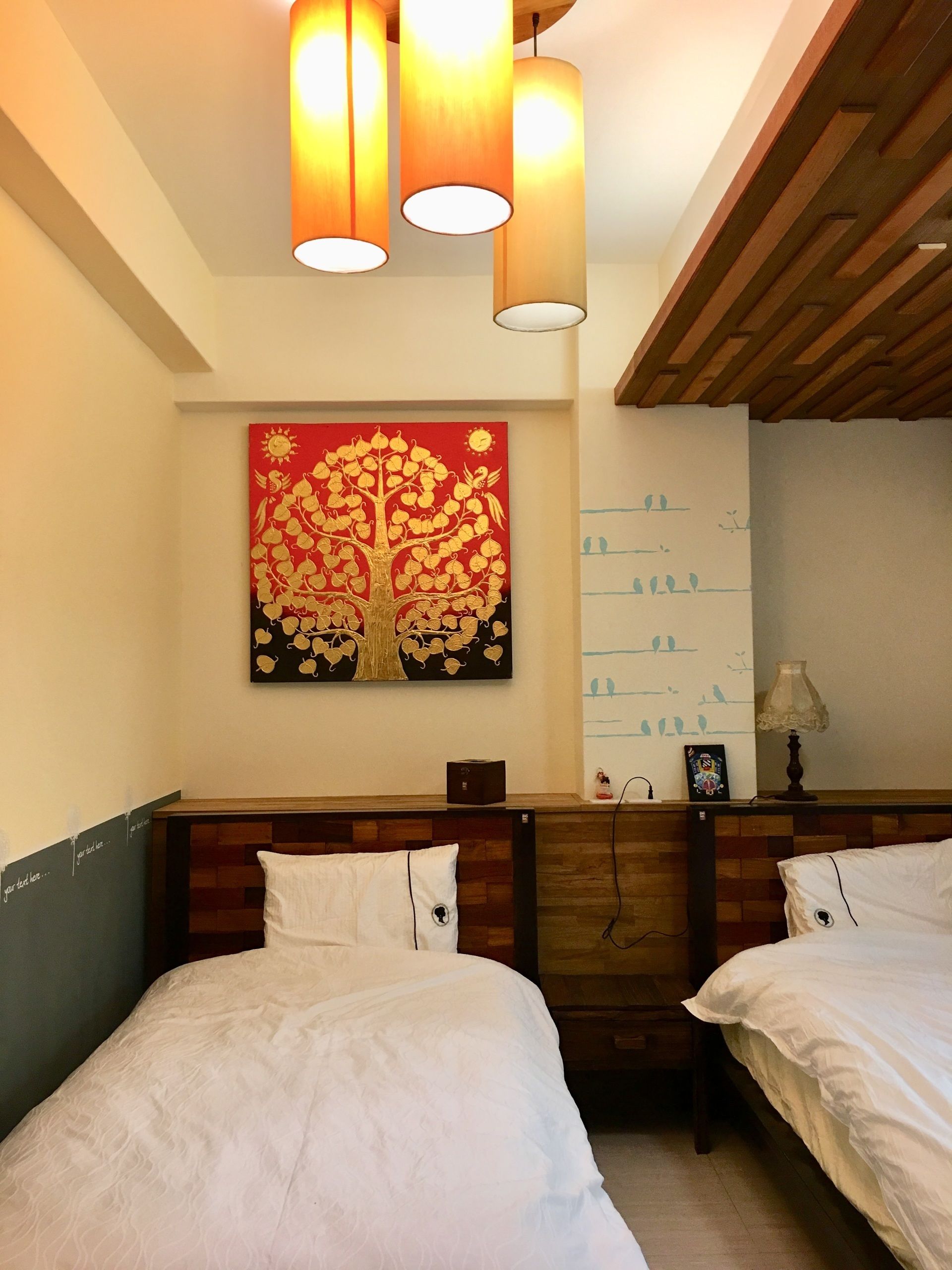 Habitación de hotel con dos camas, un cabecero de madera, un cuadro colgado en la pared con un árbol y un techo con tres lámparas colgantes.