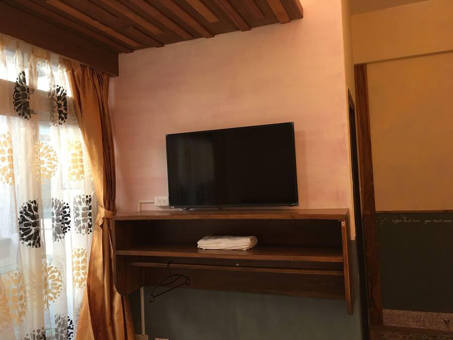 Una habitación con una televisión sobre un estante de madera, una toalla doblada en otro estante y una cortina con diseño de hojas.