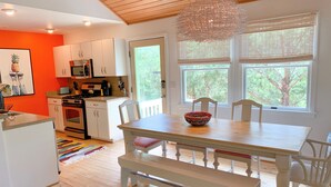 A modern kitchen with white cabinets, a wooden dining table, and a large window with a view of trees. - Fresh Fruit:  Pet Friendly, pleasant single story on private lane. (Ocracoke)