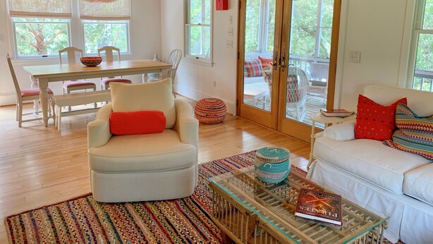 A modern living room with a white sofa, a white armchair with a red pillow, a glass coffee table with a book, and a dining area with a wooden table and chairs. - Fresh Fruit:  Pet Friendly, pleasant single story on private lane. (Ocracoke)