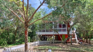 A two-story house with a red exterior and a white balcony, surrounded by trees and a wooden fence. - Fresh Fruit:  Pet Friendly, pleasant single story on private lane. (Ocracoke)