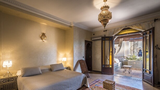 Une chambre spacieuse avec un grand lit, un coin salon comprenant un fauteuil et une petite table, et une vue sur une terrasse extérieure.