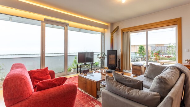 Een moderne woonkamer met een rode bank, een grijze bank en een houten salontafel. Er is een televisie en een open haard. Grote ramen bieden uitzicht op het omliggende landschap.