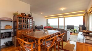 Een ruime woonkamer met een houten eettafel, zachte zitplaatsen, een kast met glazen deuren, een televisie en een groot raam voor natuurlijk licht.