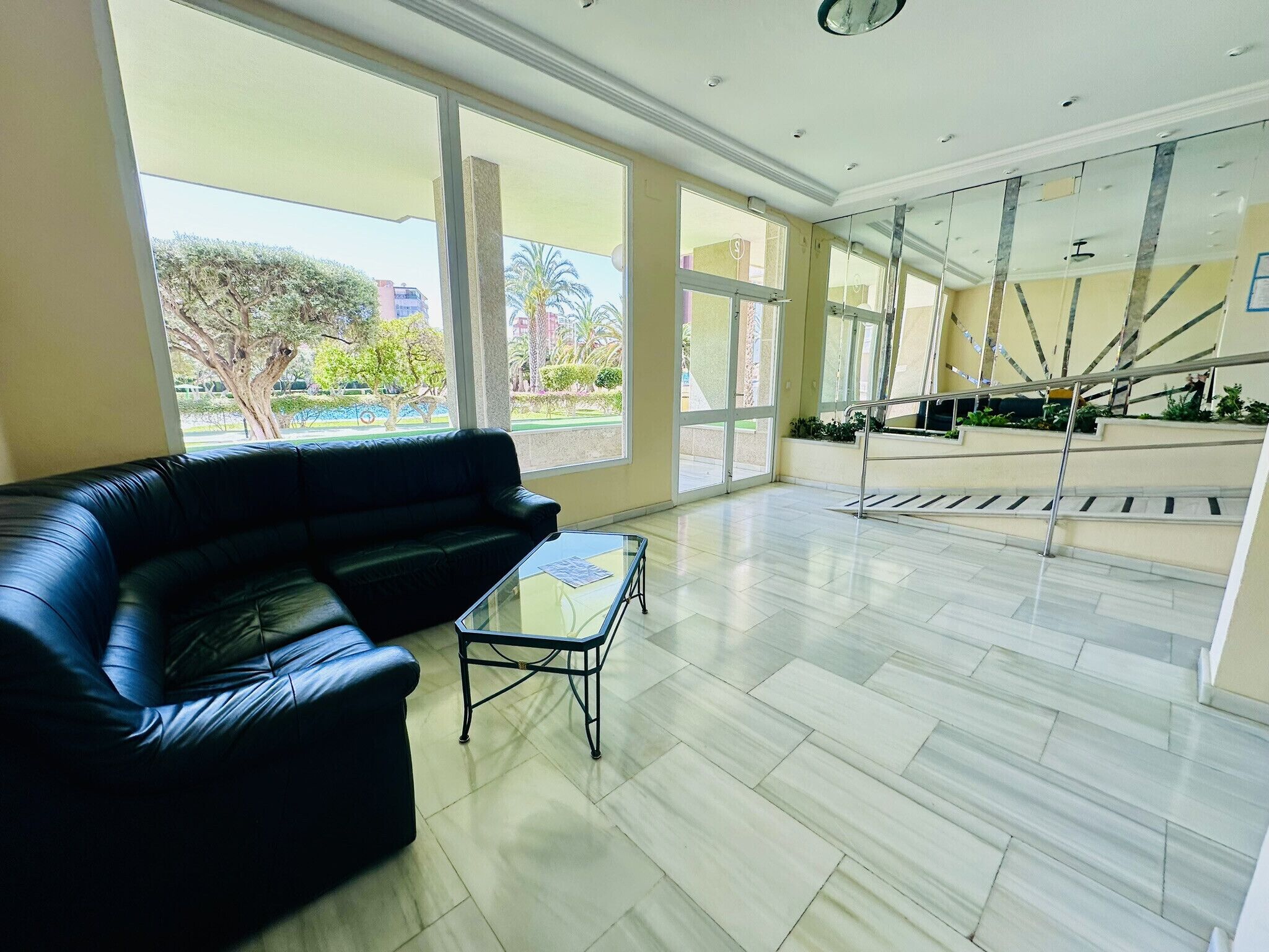 A spacious lobby with a black leather sofa, a glass coffee table, and a view of a staircase and outdoor greenery.