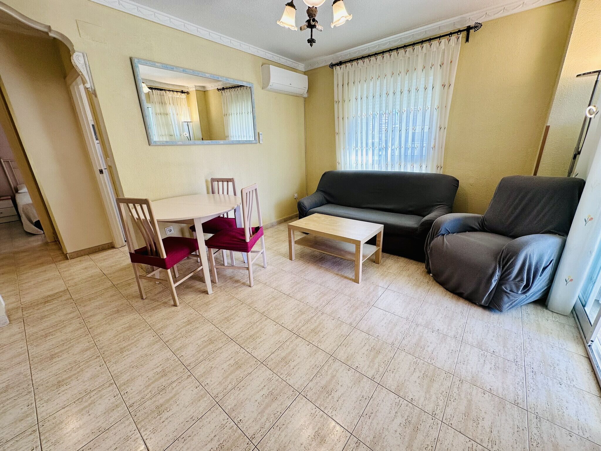 A living room with a sofa, a dining table with chairs, a small coffee table, and a large mirror on the wall.