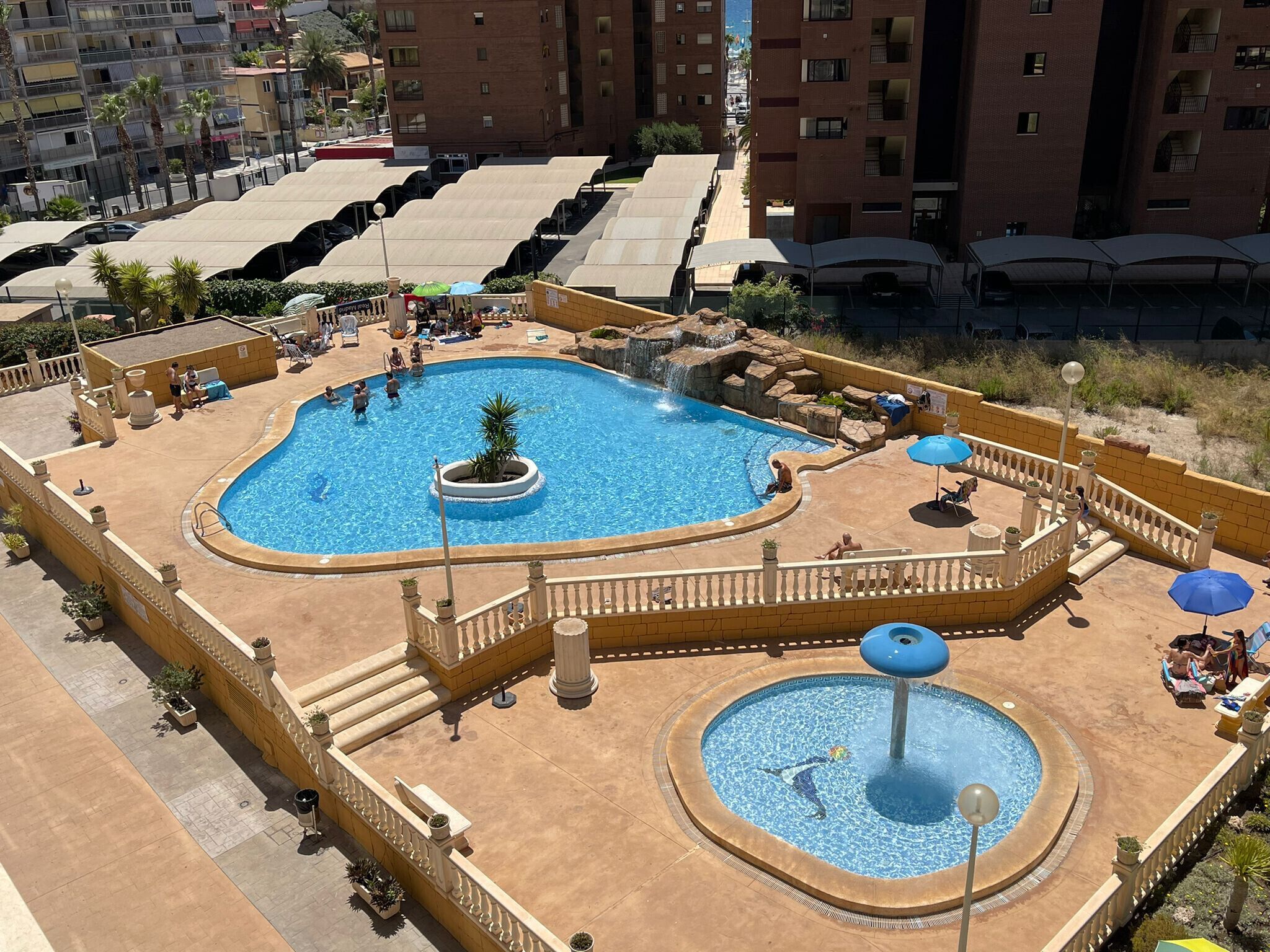 A swimming pool with a central fountain, surrounded by sun loungers and umbrellas, with a view of residential buildings in the background.