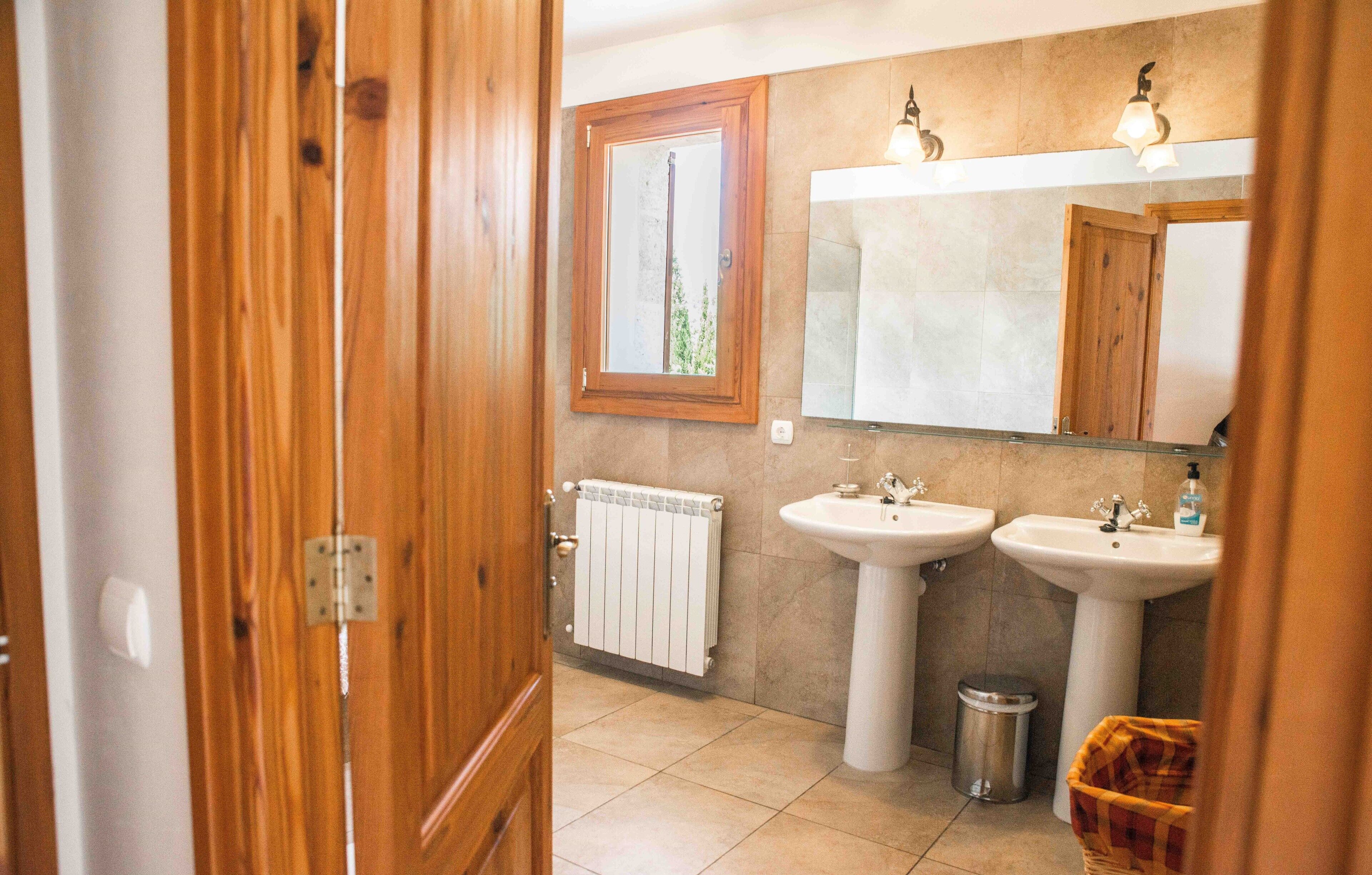 A bathroom with wooden doors, a window, two sinks, a mirror, a radiator, and a trash bin.